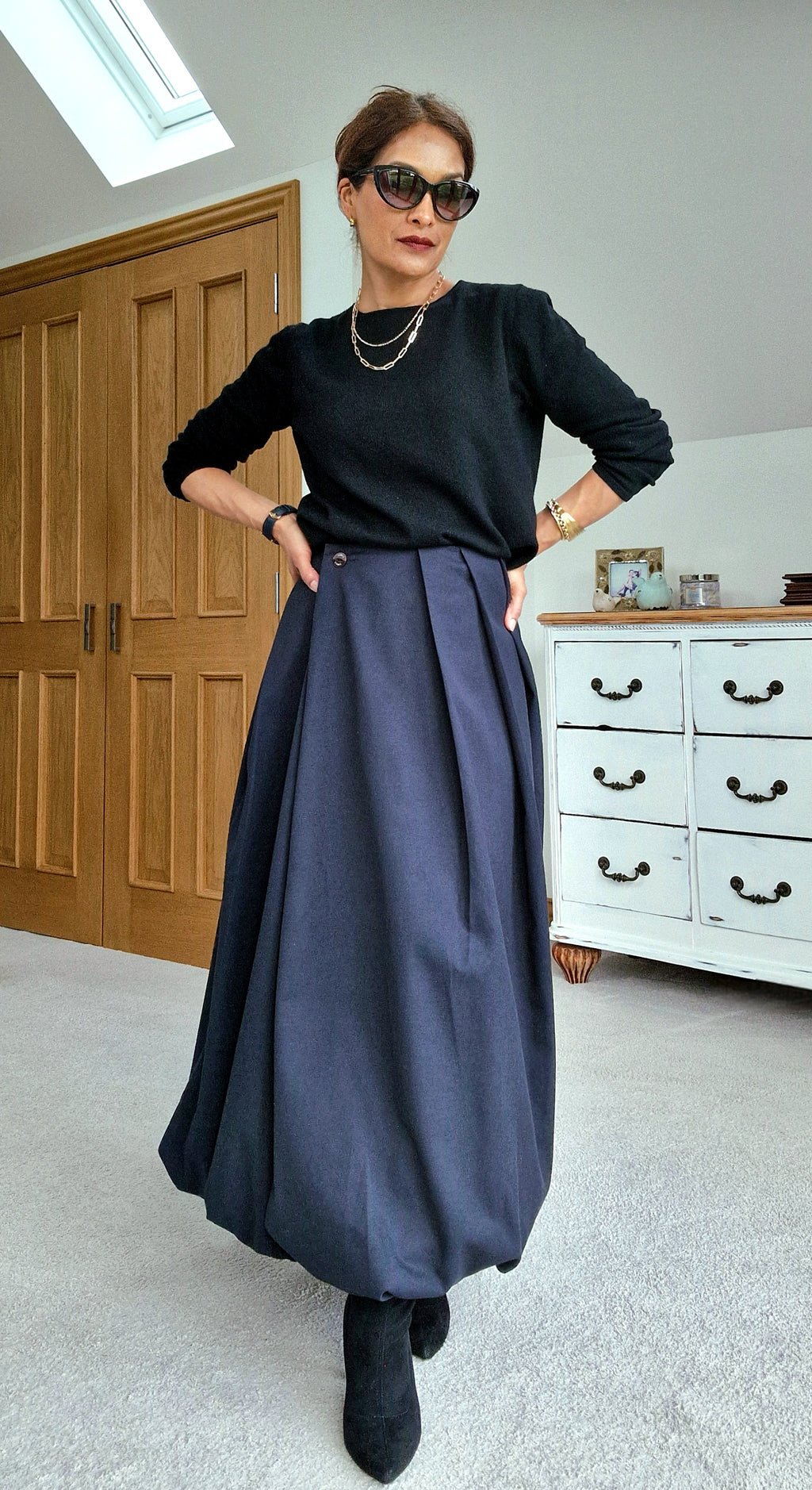 Woman wearing a black top and long blue skirt in a room with wooden cabinets and a white dresser.
