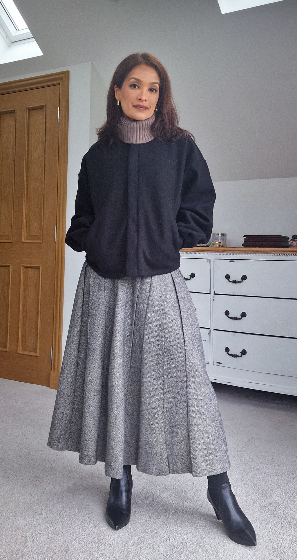 Woman wearing a black sweater and gray skirt standing in a room with a wooden door and white dresser.