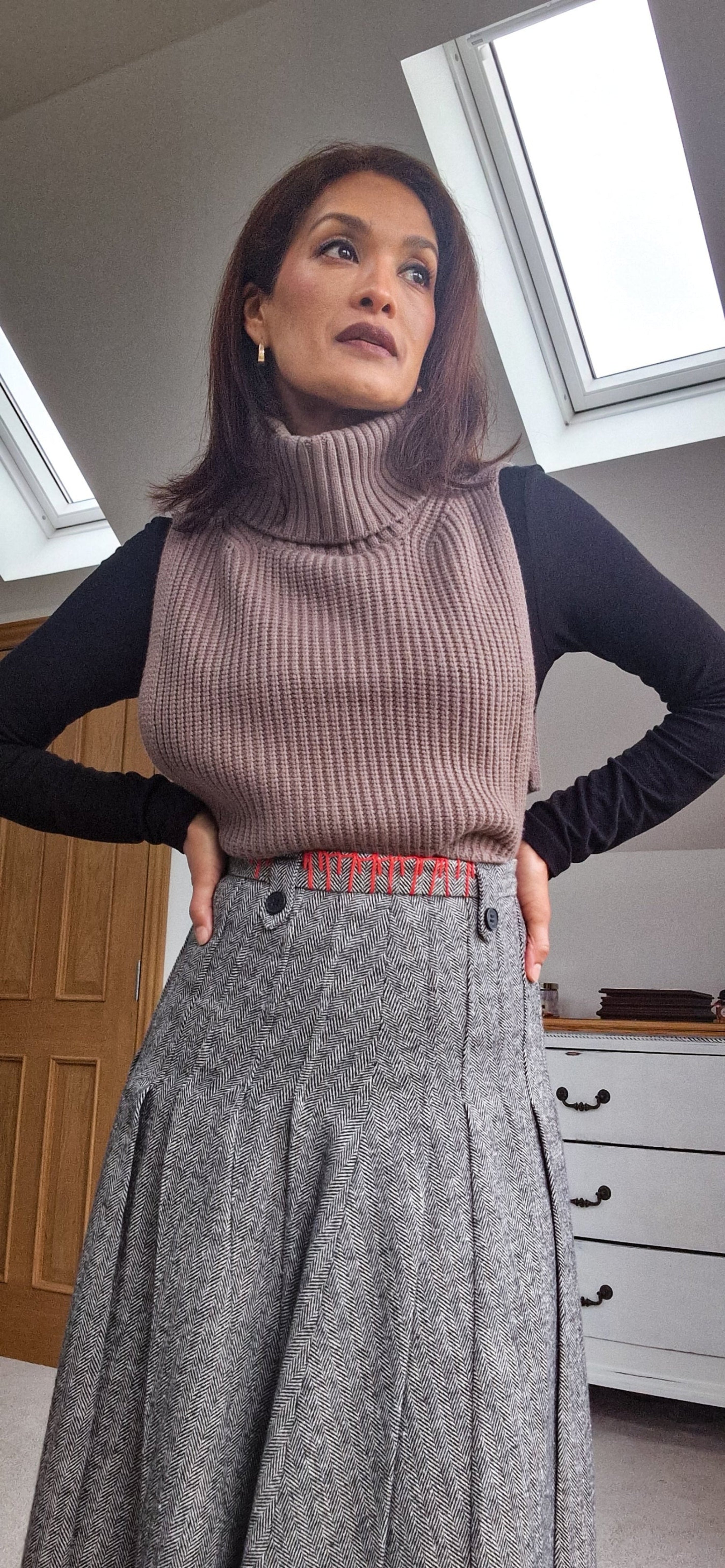 Woman wearing a brown ribbed sweater vest over a black long-sleeve shirt and gray pleated skirt indoors.