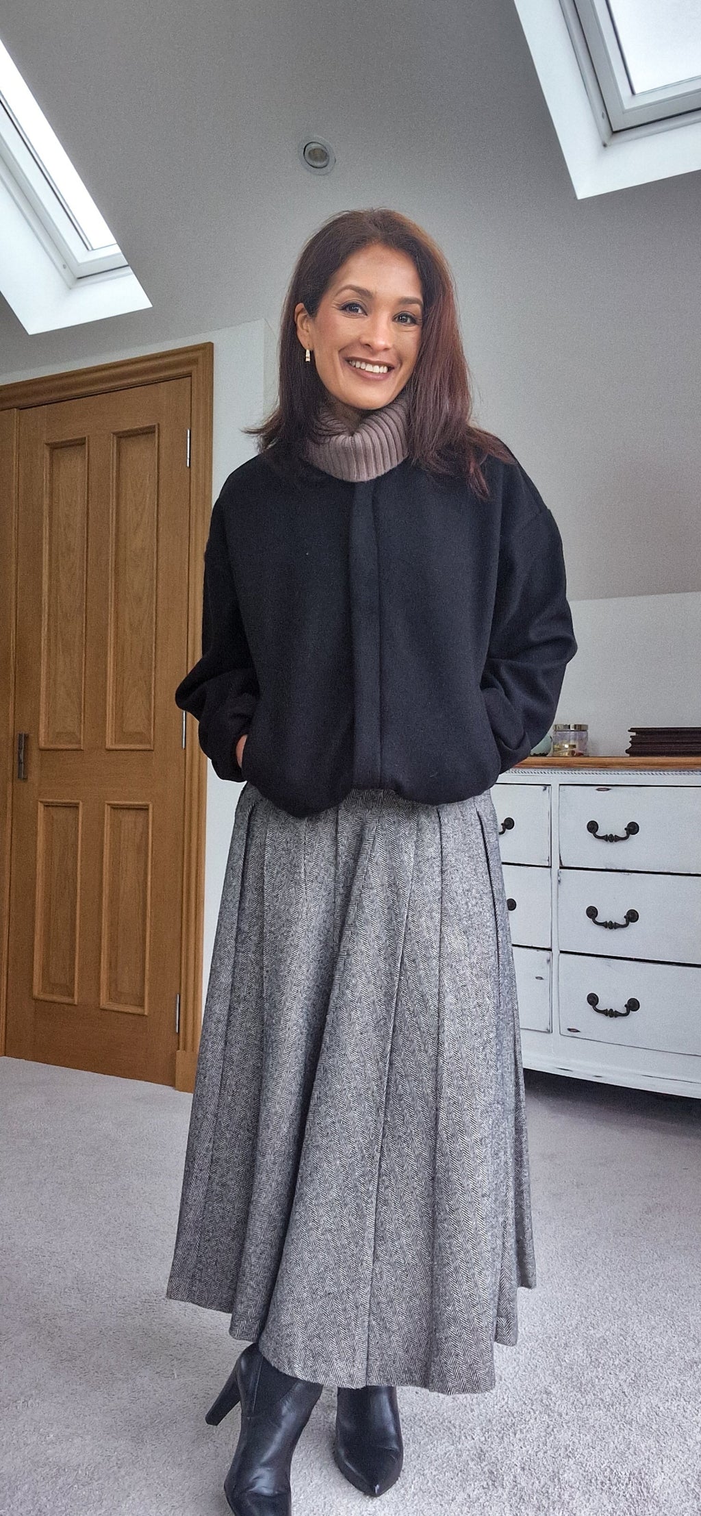 Woman wearing a black sweater and gray skirt standing in a room with wooden door and white dresser.