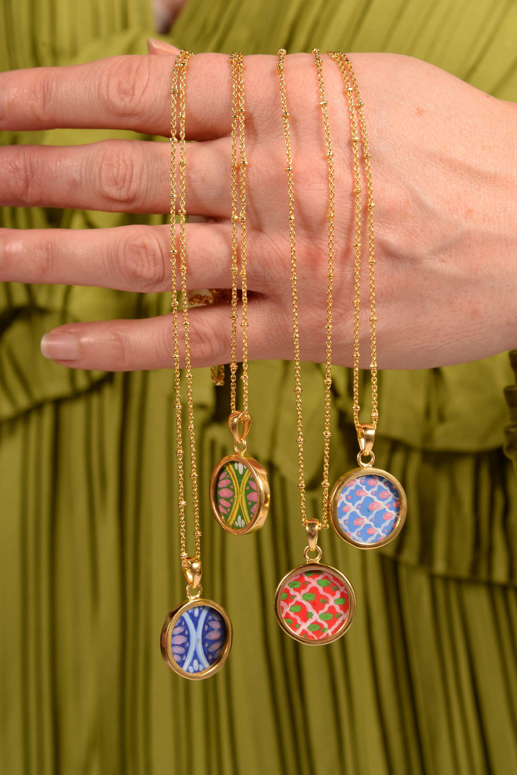 Hand holding four gold necklaces with colorful pendant designs against a green background