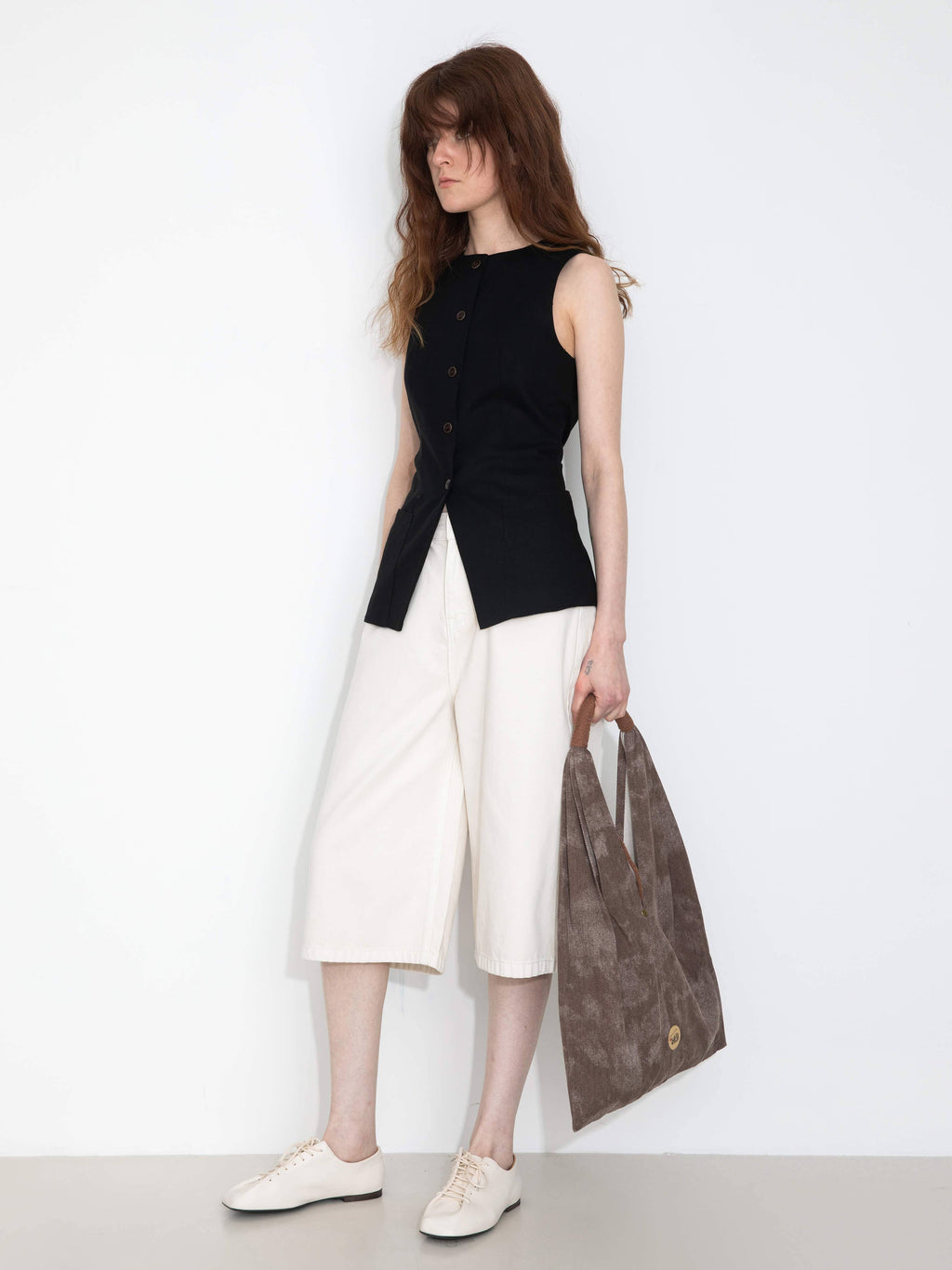 Woman wearing a black sleeveless top and white pants, holding a brown leather bag against a white background