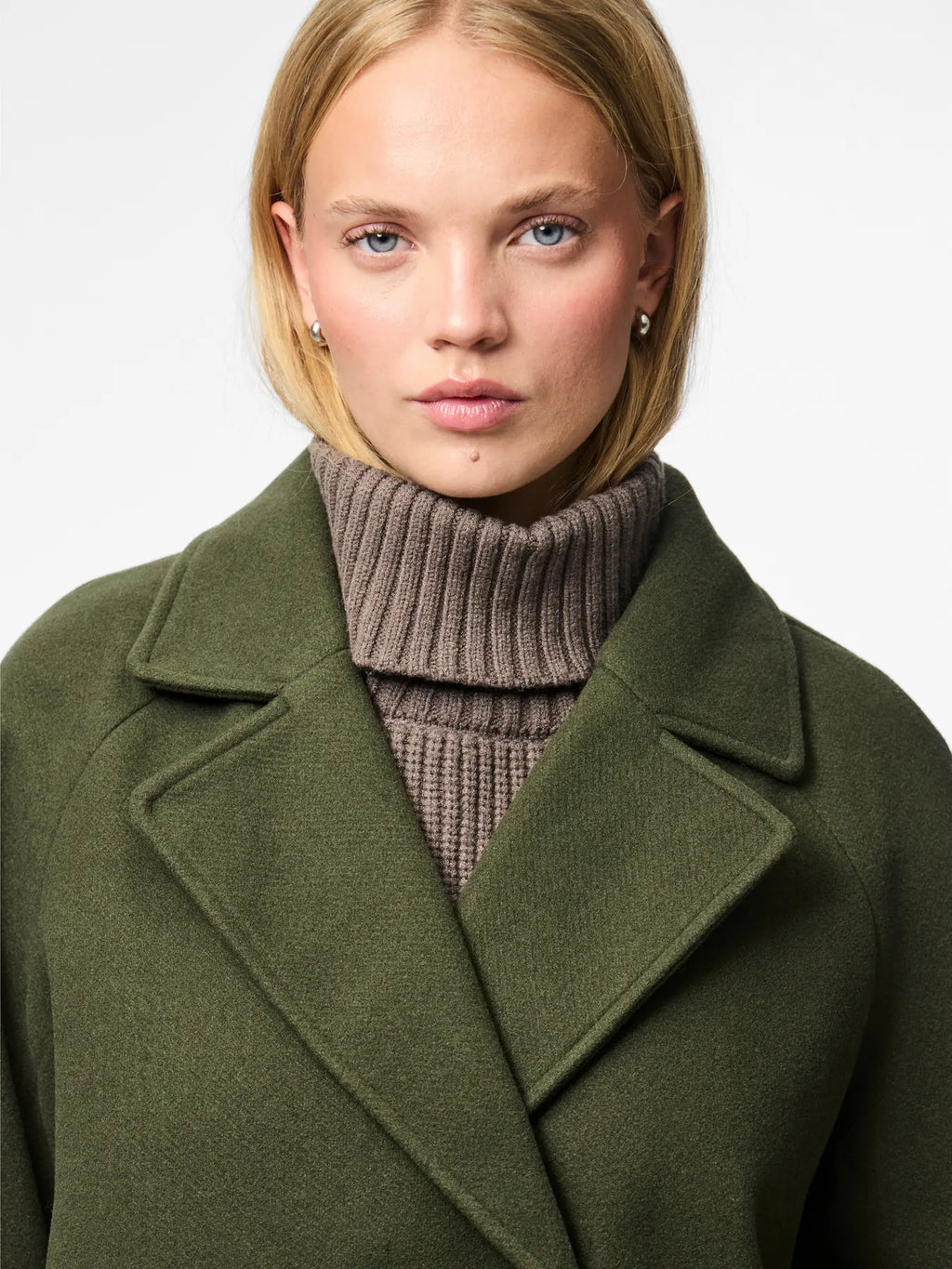 Woman wearing a green coat over a brown turtleneck sweater on a light gray background