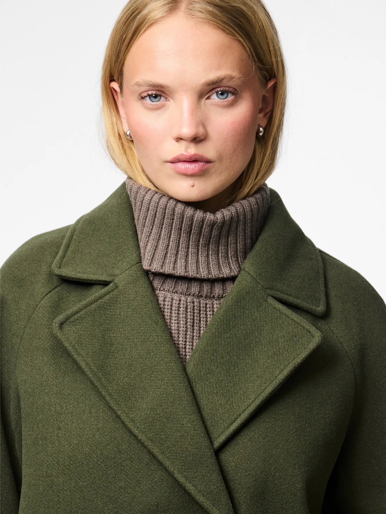 Woman wearing a green coat over a brown turtleneck sweater on a light gray background
