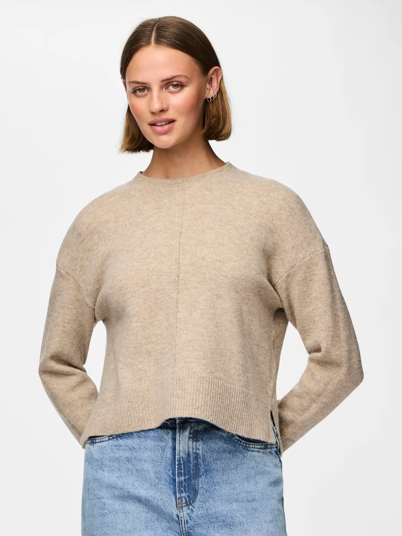 Woman wearing a beige sweater and blue jeans on a light gray background