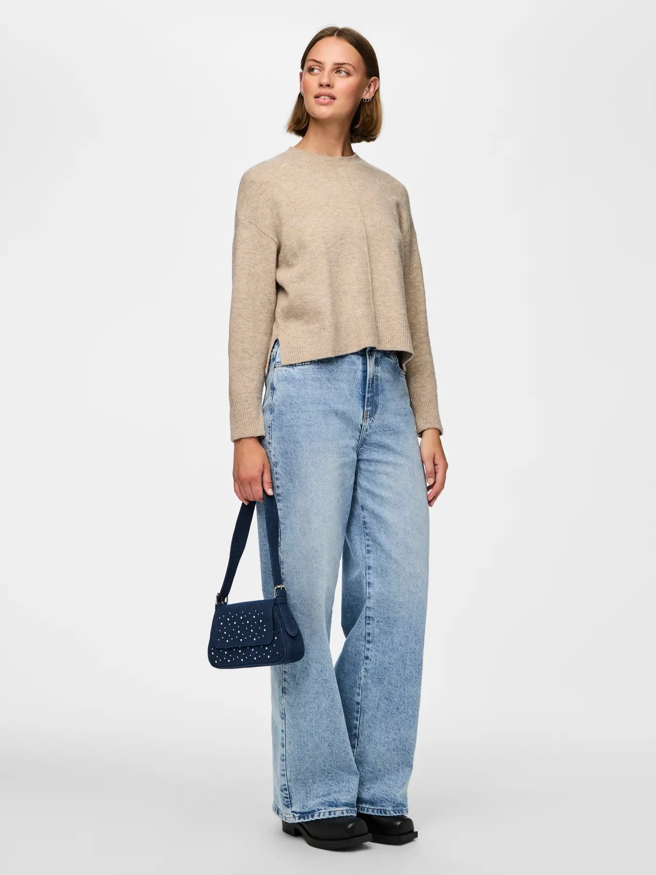 Woman wearing a beige sweater and blue jeans holding a black handbag on a white background