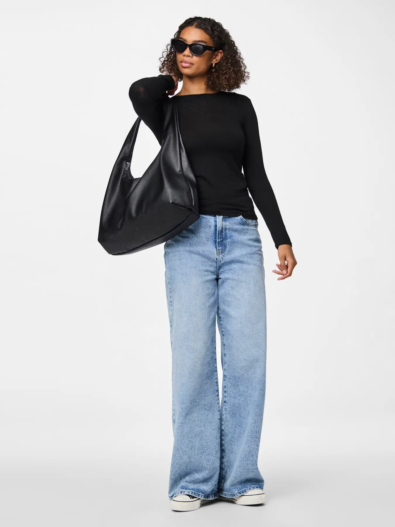 Woman holding a black leather handbag wearing sunglasses, a black top, and light blue jeans on a white background
