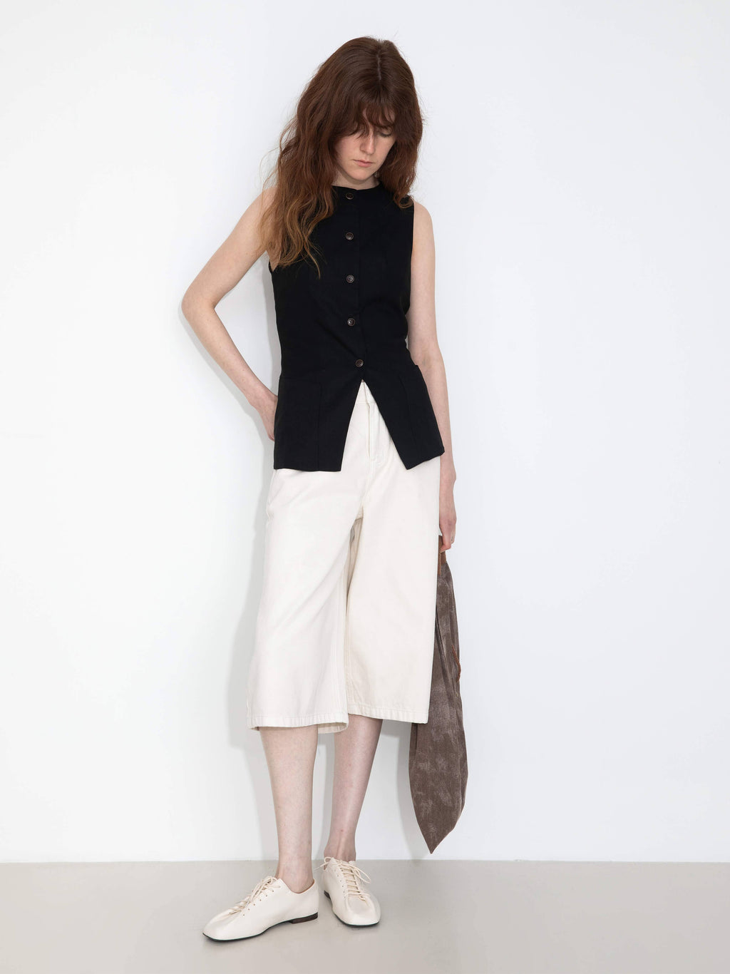 Woman wearing a black sleeveless top and white pants with a long dark scarf, standing against a plain background.