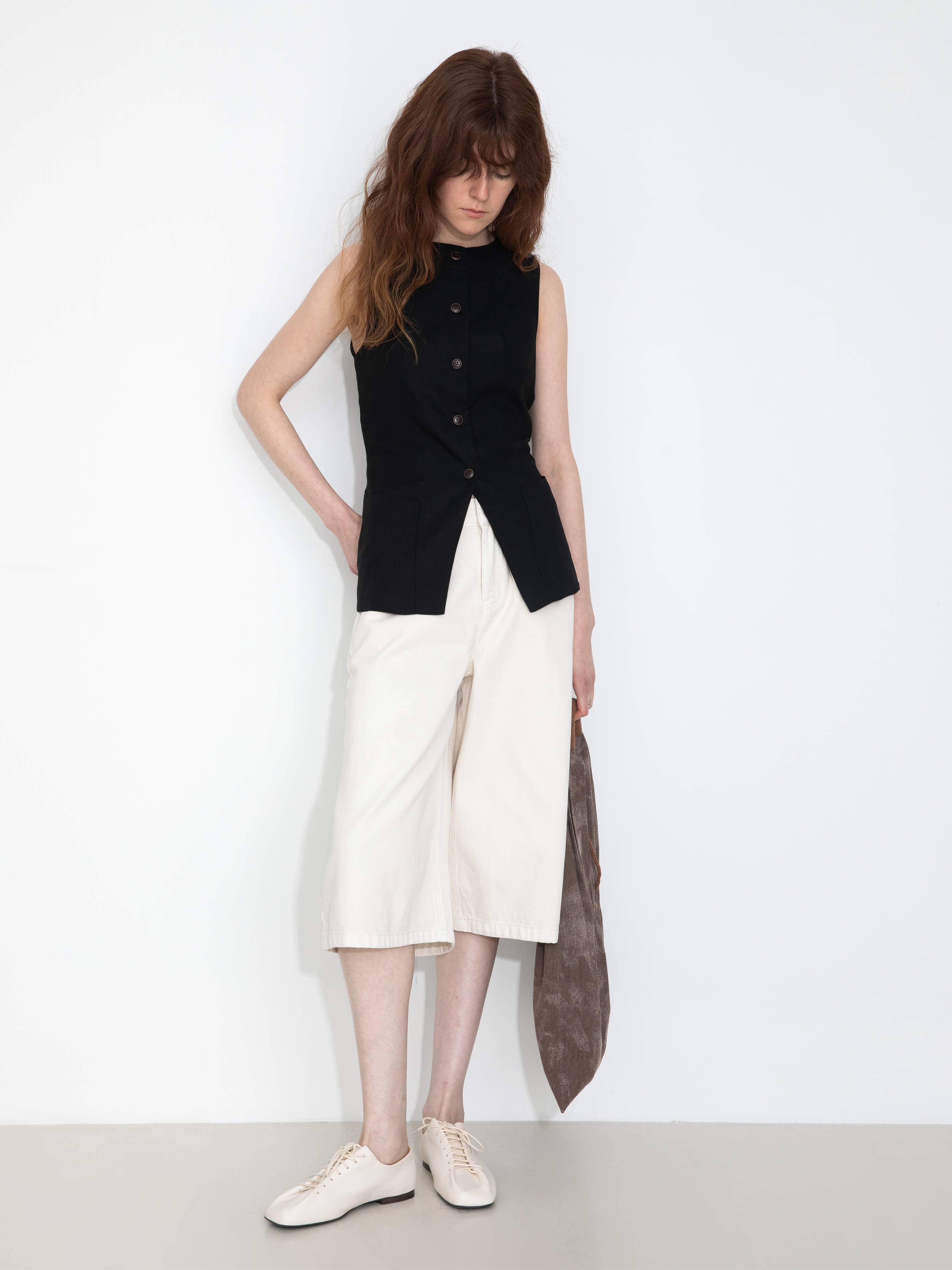 Woman wearing a black sleeveless top and white pants with a long dark scarf, standing against a plain background.